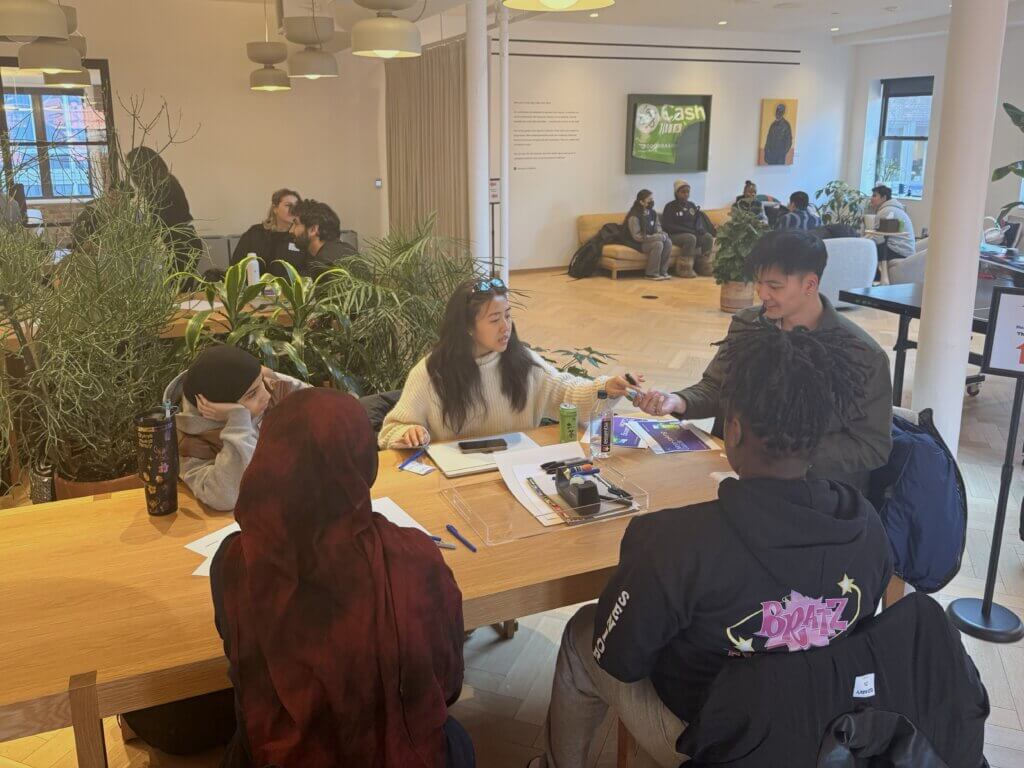 Four students sit around a table with notebooks and laptops, engaged in discussion in a modern, well-lit common area with other people in the background.