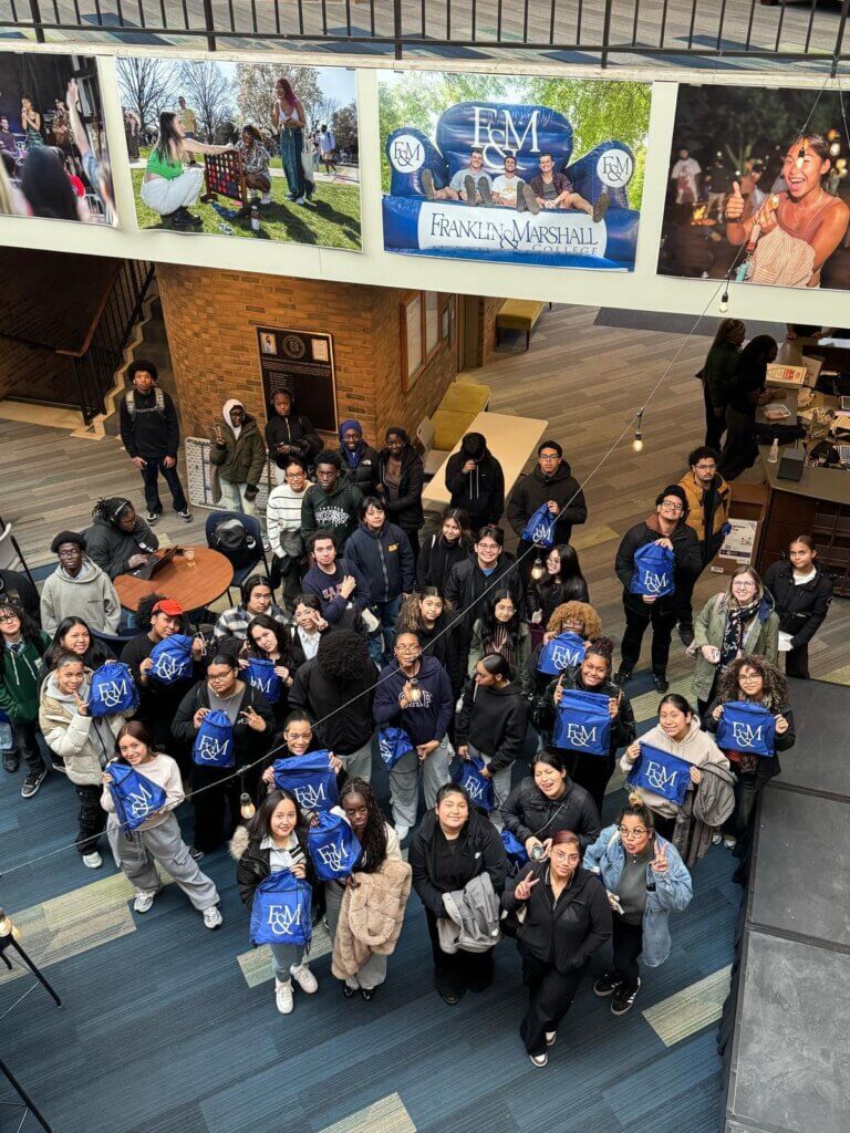 A group of students poses for a photo indoors, many wearing Franklin & Marshall sweatshirts, with college banners and photos proudly displayed above them.