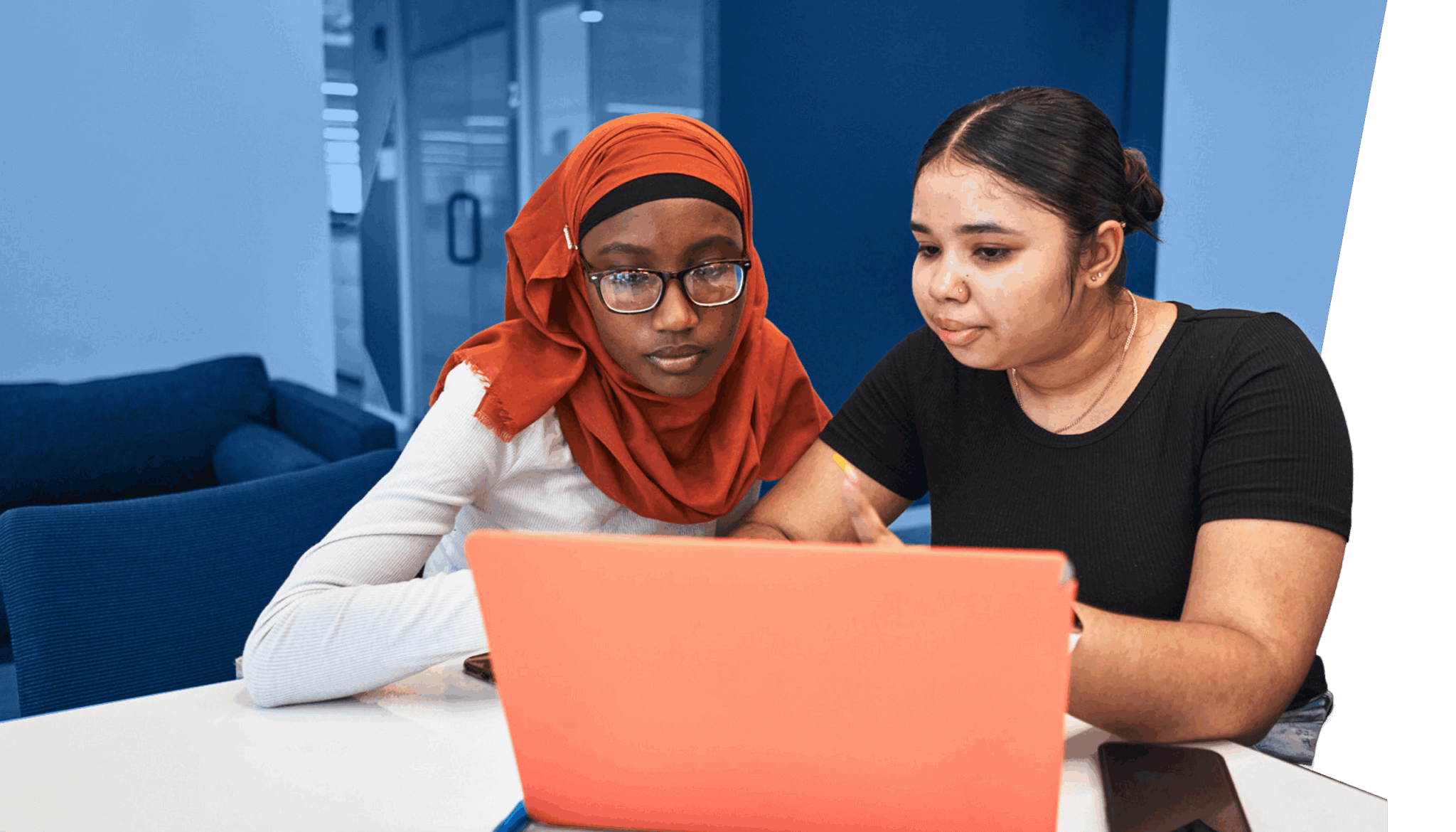 Two women sit at a table working together on a laptop in a modern office setting, focusing on college persistence and graduation support, with a smartphone and pen on the table.