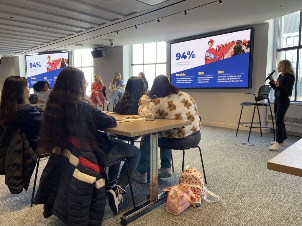 A group of people sits at tables listening to a speaker present data slides, including information about the SUNY McConney EOP award, on two large monitors in a modern conference room with large windows.