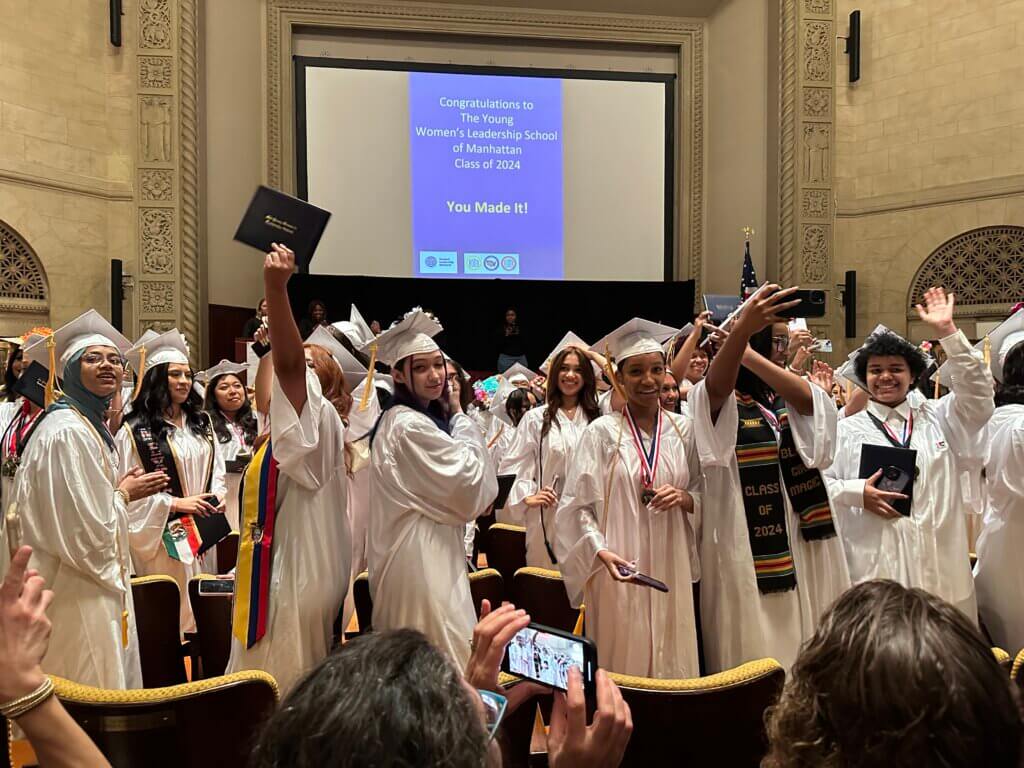 A group of students in white graduation gowns and caps celebrate at a commencement ceremony, holding diplomas and smiling in an auditorium.