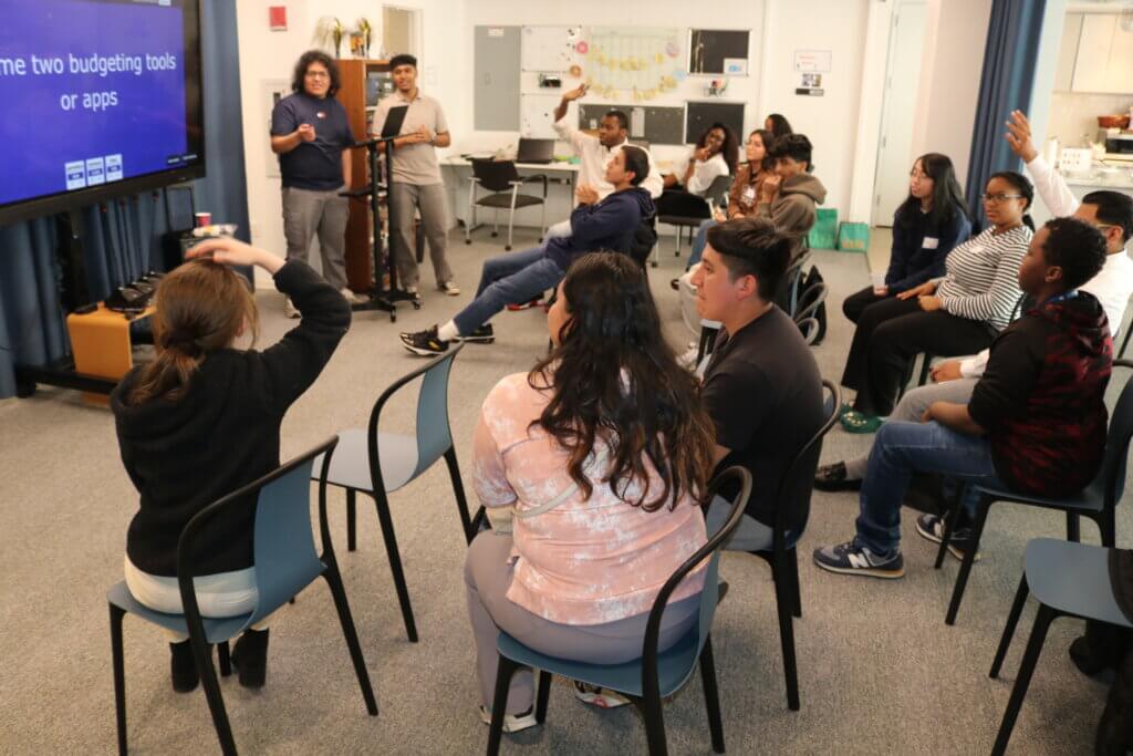 A group of students sit facing a screen displaying a budgeting question, some raising their hands to ask FAQs, while two presenters—CBI Alumni—lead the session at the front of the room.