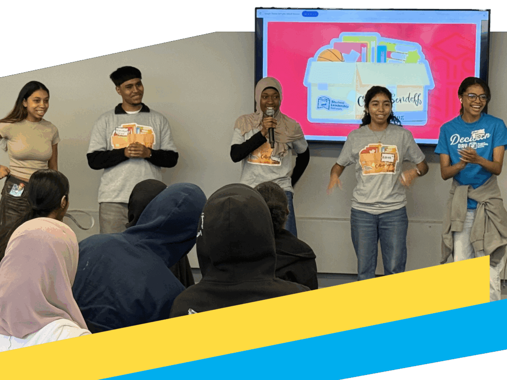 Five young people stand in front of an audience in a classroom, presenting near a screen displaying colorful folders and documents.