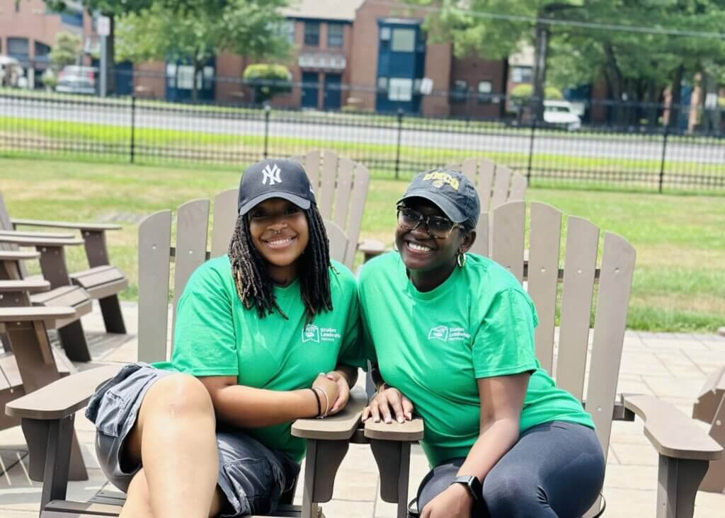 Two women, showcasing student leadership, wear green T-shirts and black caps as they sit together on wooden chairs outdoors, smiling at the camera.