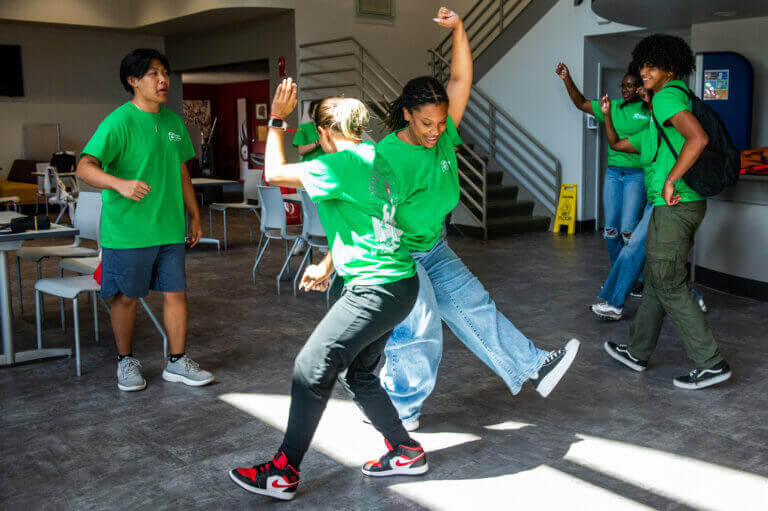 Five people in green shirts, showing student leadership, interact and playfully dance in a bright, modern room with tables, chairs, and a staircase in the background.