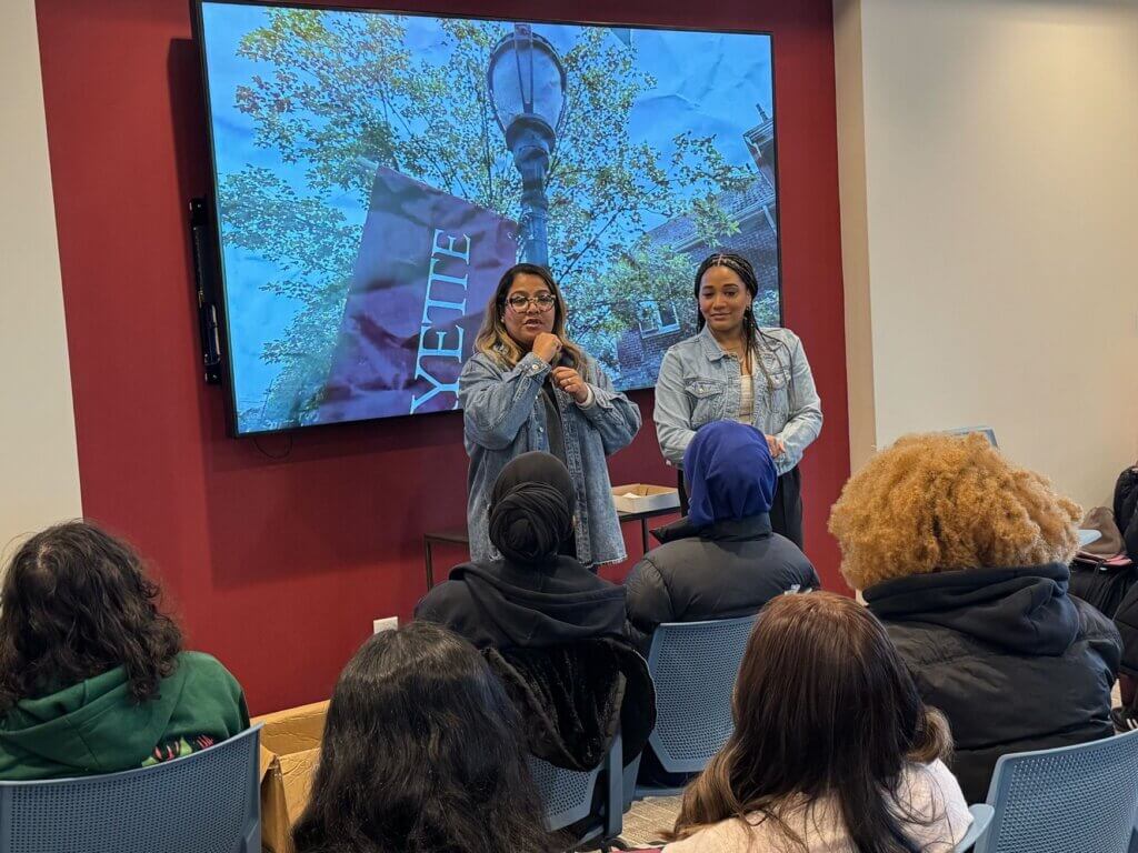 Two women stand and speak in front of an audience, with a large screen behind them displaying an outdoor image and the word Lafayette