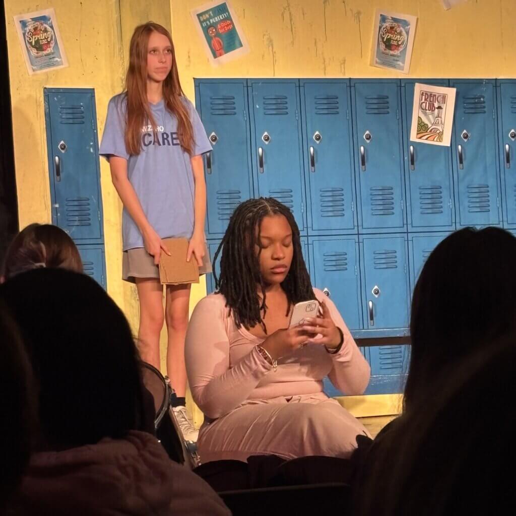 Two teenage girls are on stage in front of blue lockers, embodying student leadership; one stands holding a notebook, while the other sits with her phone. Audience members are visible in the foreground.