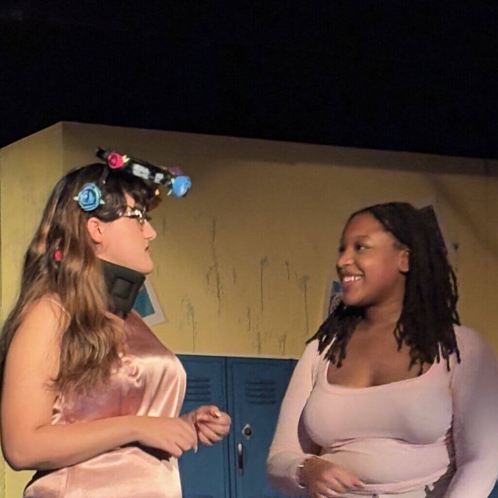 Two women stand and talk in front of blue lockers. One wears a neck brace and a headpiece with flowers; the other, showcasing student leadership, wears a light pink top and smiles.