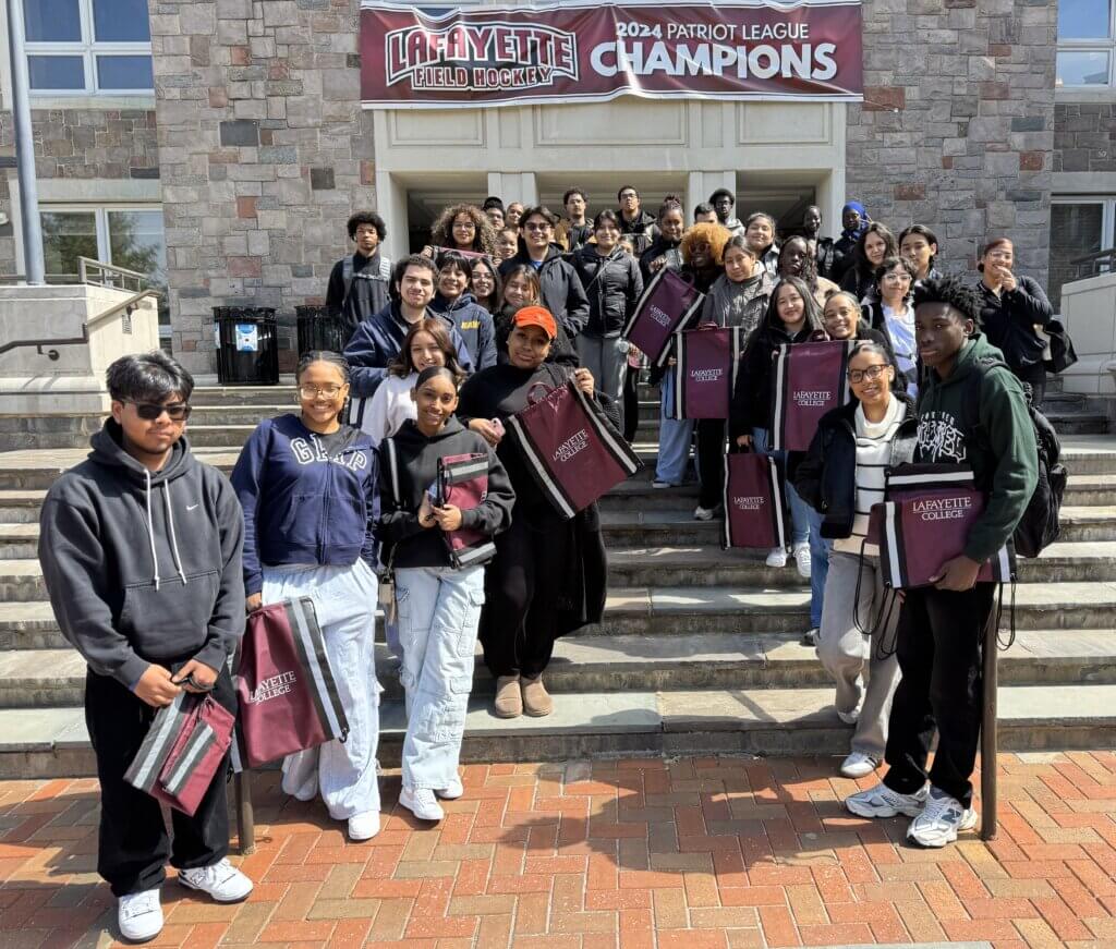 A group of students stands on the steps outside a building with a “Lafayette Field Hockey 2024 Patriot League Champions” banner, proudly displaying maroon Lafayette tote bags and a SUNY McConney EOP award.