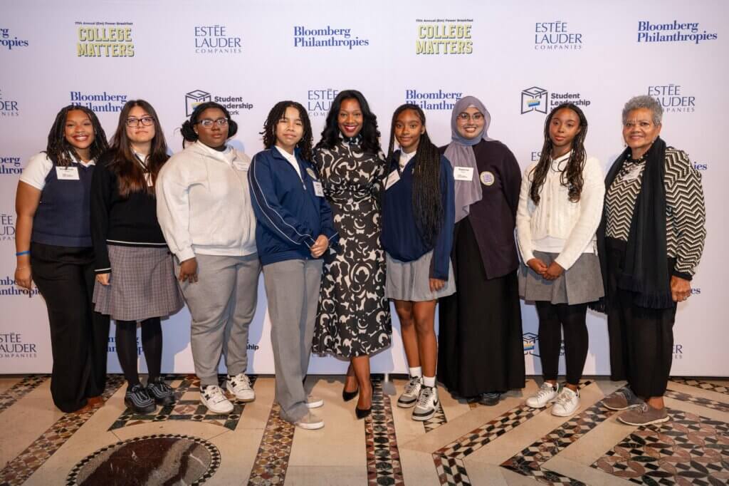 A group of eight women and girls, showcasing student leadership, stand together and smile for a photo at an event with a backdrop featuring various sponsor logos.