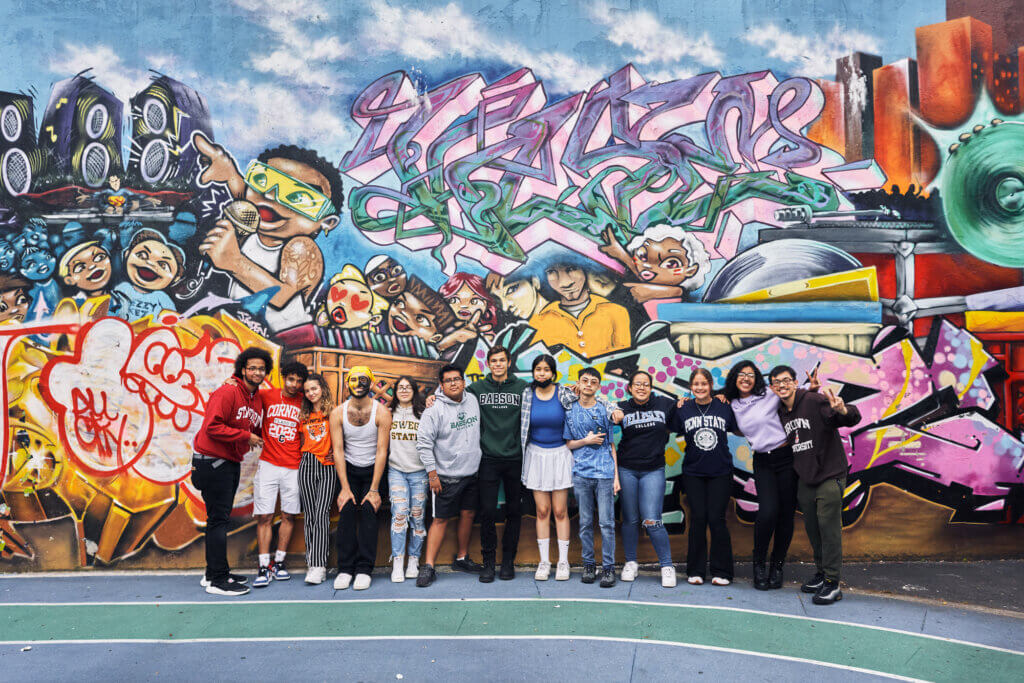 A group of teenagers stands in front of a colorful, graffiti-covered wall on an outdoor basketball court.