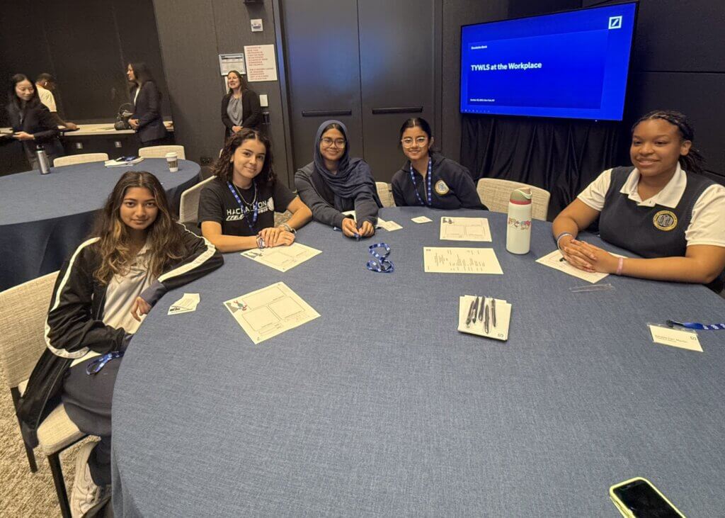 Five young women sit at a round table with papers, pens, and a water bottle, attending a TWHS at the Workplace event focused on student leadership in a conference room.