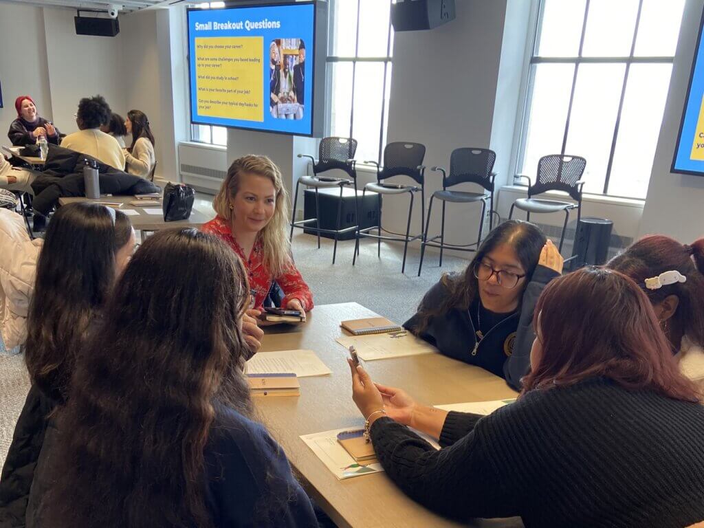 A group of people sit around a table having a discussion, with a presentation slide displaying small breakout questions and the SUNY McConney EOP award visible in the background.