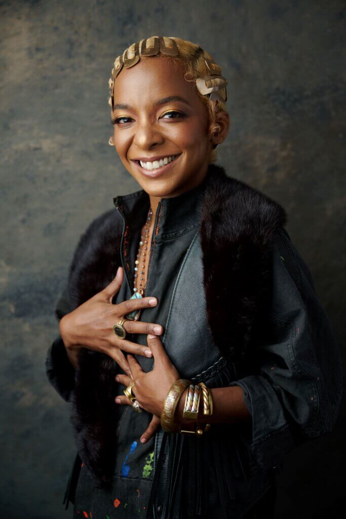 Alumna Kara Young, with short blonde hair clips, wearing a dark outfit with a fur vest, layered necklaces, rings, and bracelets, smiles at the camera against a textured background.