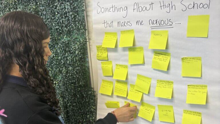 A student places a sticky note on a board labeled Something About High School that makes me nervous
