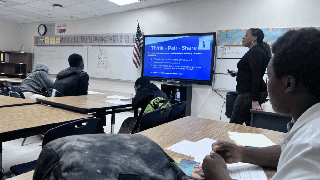 Carolina stands by a screen displaying instructions for a Think-Pair-Share activity while students sit at desks and take notes in a classroom.