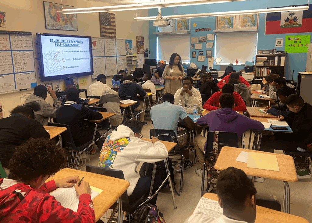 A classroom full of students sits at desks working on assignments while a teacher stands at the center; a screen displays Study Skills & Habits Self-Assessment.
