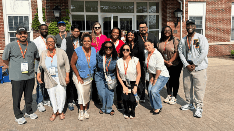 A group of sixteen adults posing and smiling outside a brick building, all wearing orange lanyards, with name badges visible on some individuals.
