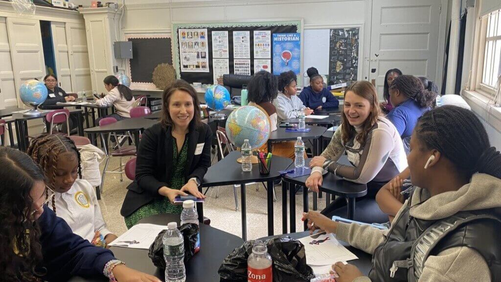 Students and two adults sit in groups at desks in a classroom with globes, papers, and drinks, engaging in discussion