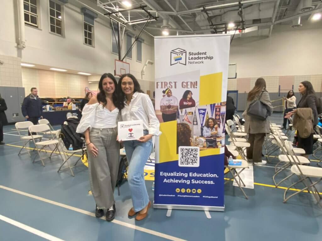 Two women stand and smile in a gymnasium, one holding a certificate, next to a Student Leadership Network banner