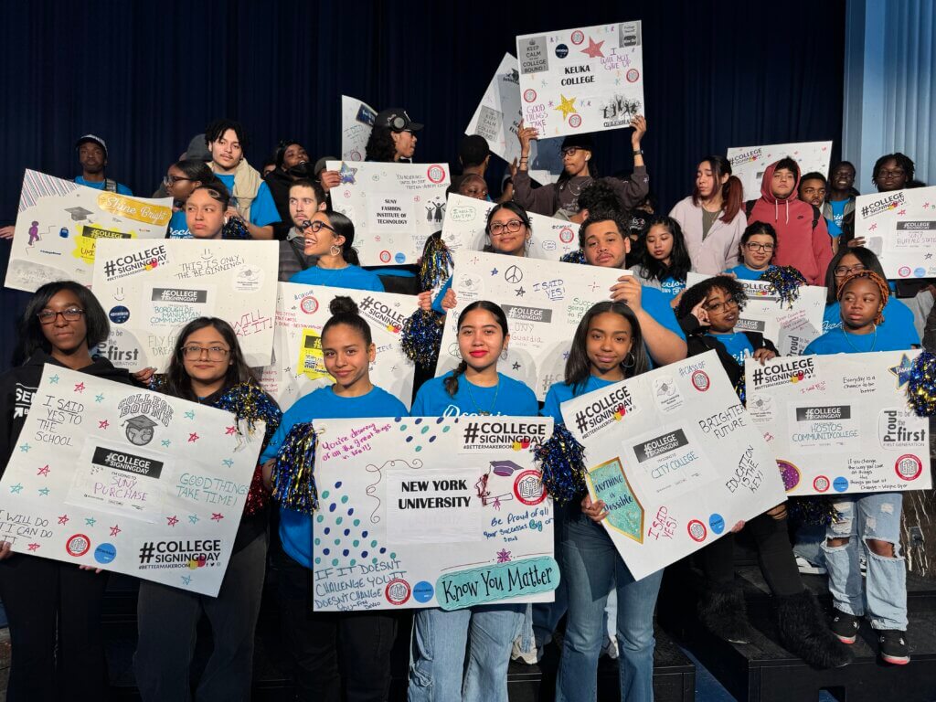 A group of students stand together holding decorated college signing posters, some wearing blue T-shirts that read #CollegeSigningDay.