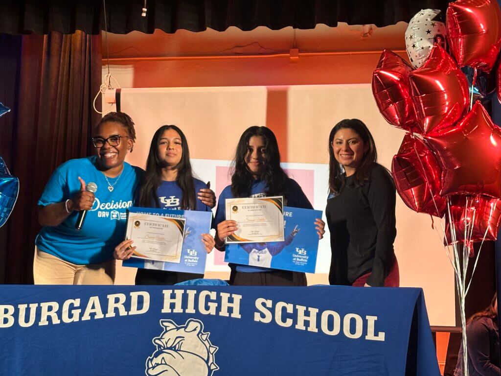 Four women stand behind a table with a Burgard High School cloth; two hold certificates and folders, while one gives a thumbs up. Balloons and a projector screen are in the background.