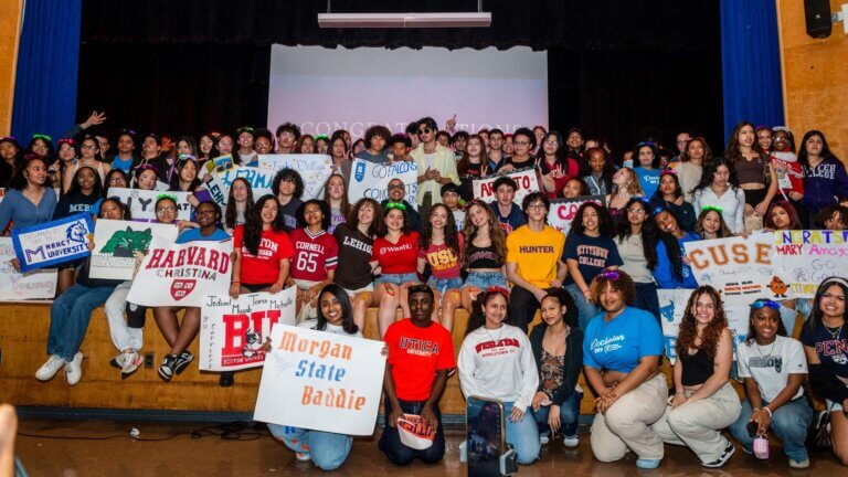 Large group of students posing on a stage, holding college signs and wearing shirts from various universities and colleges, celebrating college decisions.