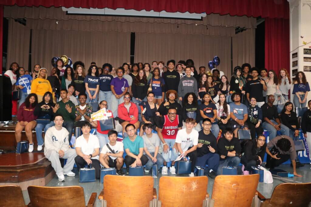 A large group of students pose on an auditorium stage, many wearing college shirts and holding gift bags, in front of a red curtain and tiered seating.
