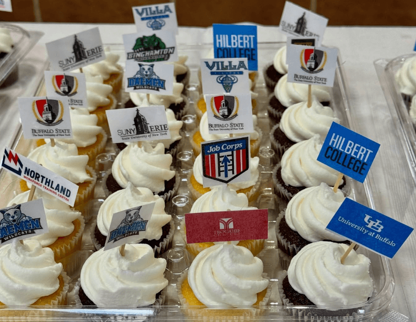 A tray of cupcakes topped with small flags displaying various college and organization logos, including Hilbert College, SUNY Erie, and Buffalo State.