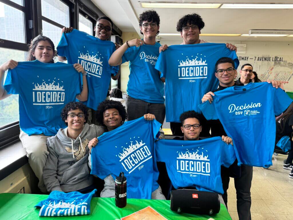 A group of students indoors hold up blue I Decided T-shirts and smile at the camera, with others wearing matching shirts.