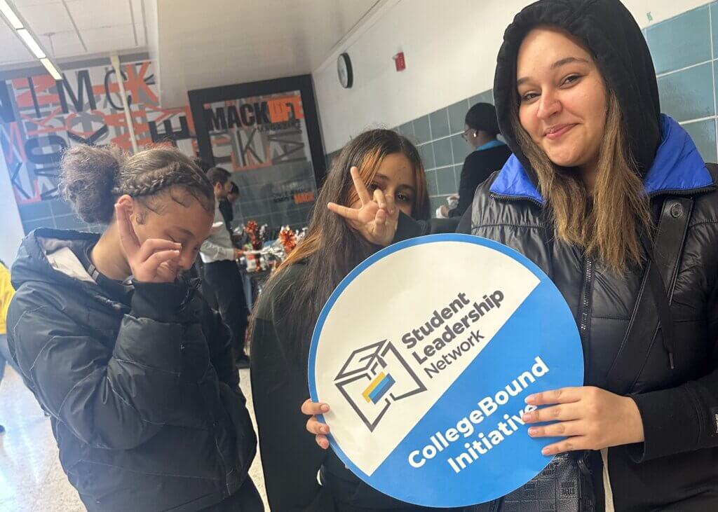 Three students stand in a school hallway holding a round sign that reads “Student Leadership Network CollegeBound Initiative.”.