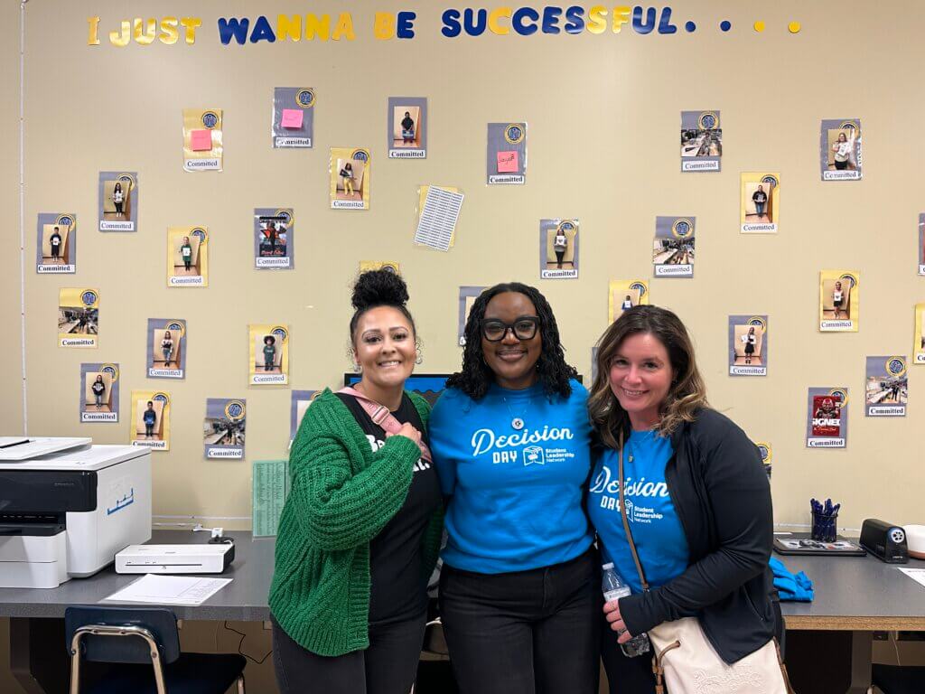 Three women stand smiling in a classroom decorated with student photos and the phrase I just wanna be successful on the wall. Two of the women wear blue Decision Day shirts.