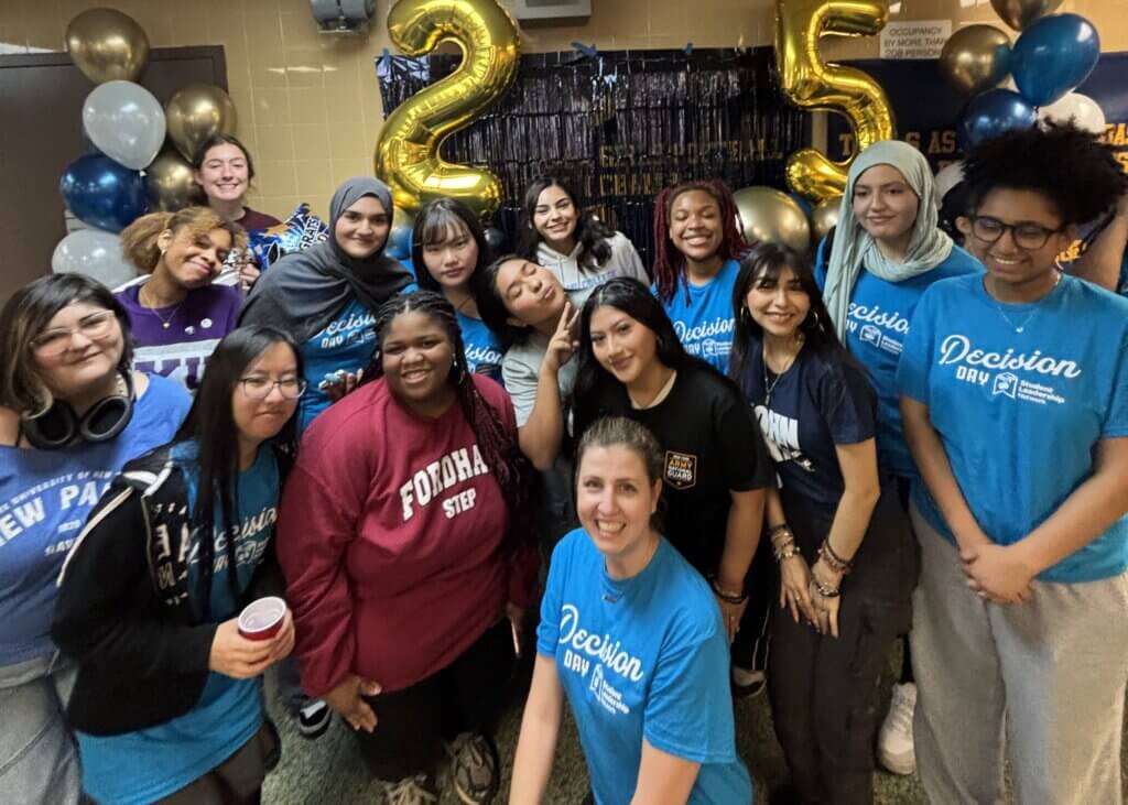 A group of people, many wearing Decision Day shirts, pose together indoors in front of gold balloon numbers 2 and 5 and decorative balloons.