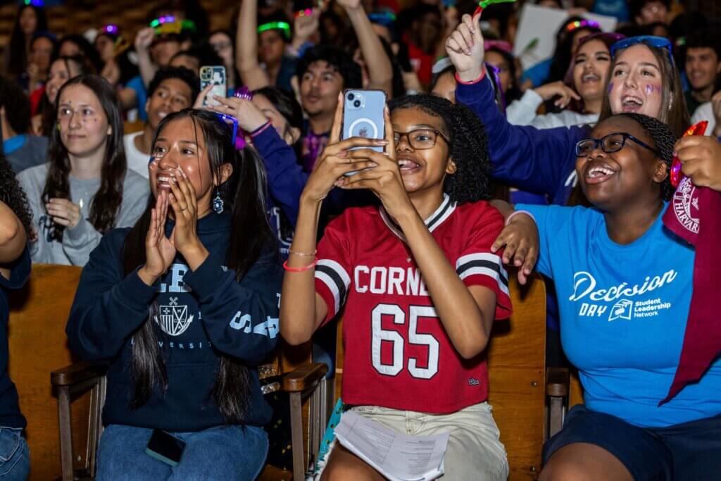 A group of students sitting in an auditorium cheer and smile, with one student taking a photo on a phone and others clapping and wearing college-themed shirts.