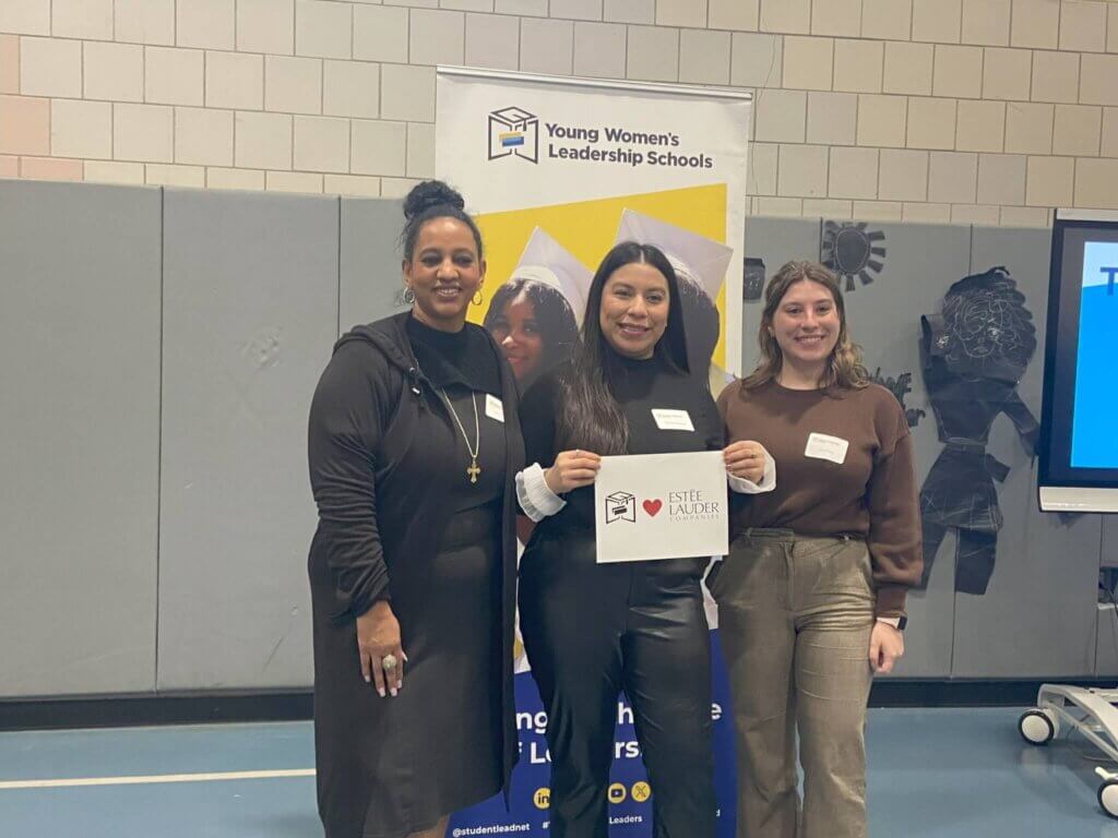 Three women stand in front of a Young Women’s Leadership Schools banner; the center woman holds a sign with the Estée Lauder logo and heart.
