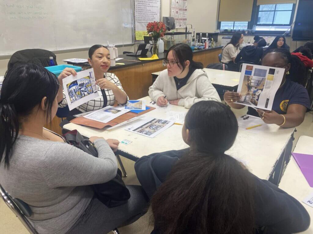 Four students sit around a classroom table, discussing and holding printed images