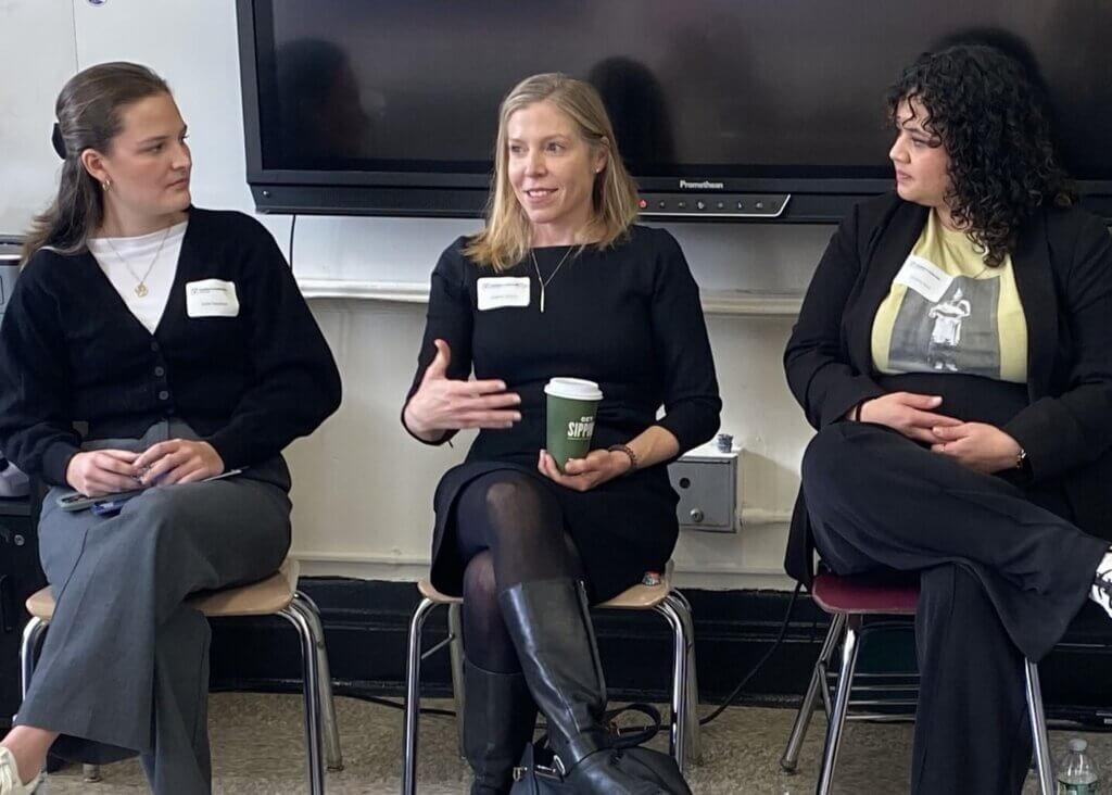 Three women sit on chairs, engaged in conversation. The woman in the center gestures with her hand and holds a coffee cup, while the other two listen attentively.