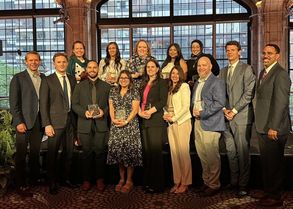 A group of thirteen people, some holding glass awards, pose together on a stage in a formal indoor setting with large windows in the background.