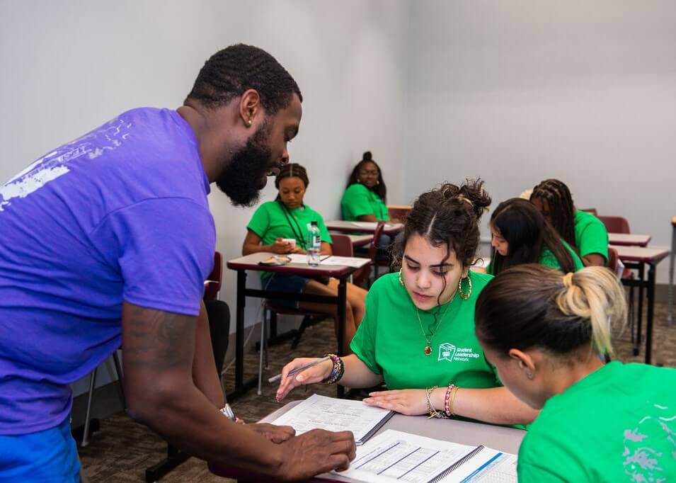 A teacher in a purple shirt assists students wearing green shirts with their work at desks in a classroom. Other students are seated and working in the background.