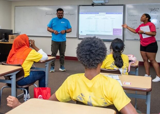 Two instructors stand at the front of a classroom presenting to students, with a projected spreadsheet on the whiteboard and students seated at desks facing them.