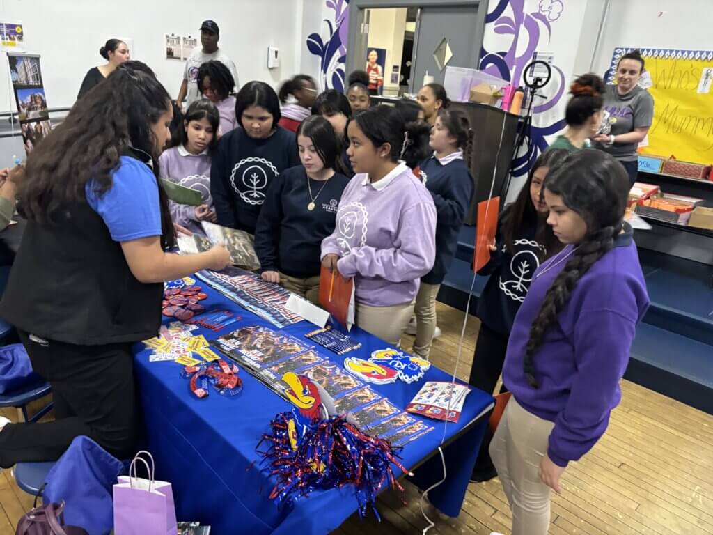 A group of middle school students gathers around a table with college promotional materials and merchandise
