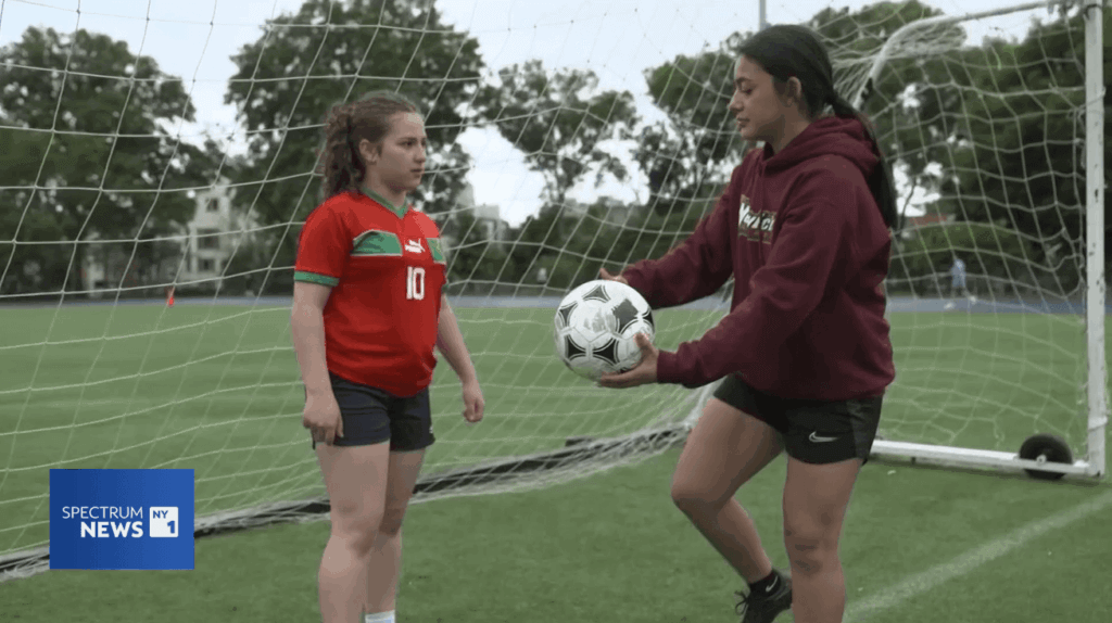 Student Dahna in a maroon hoodie hands a soccer ball to a younger student by the goal post