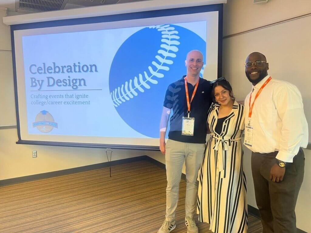 Three people stand in front of a presentation screen displaying “Celebration By Design” with a baseball graphic in a conference room.