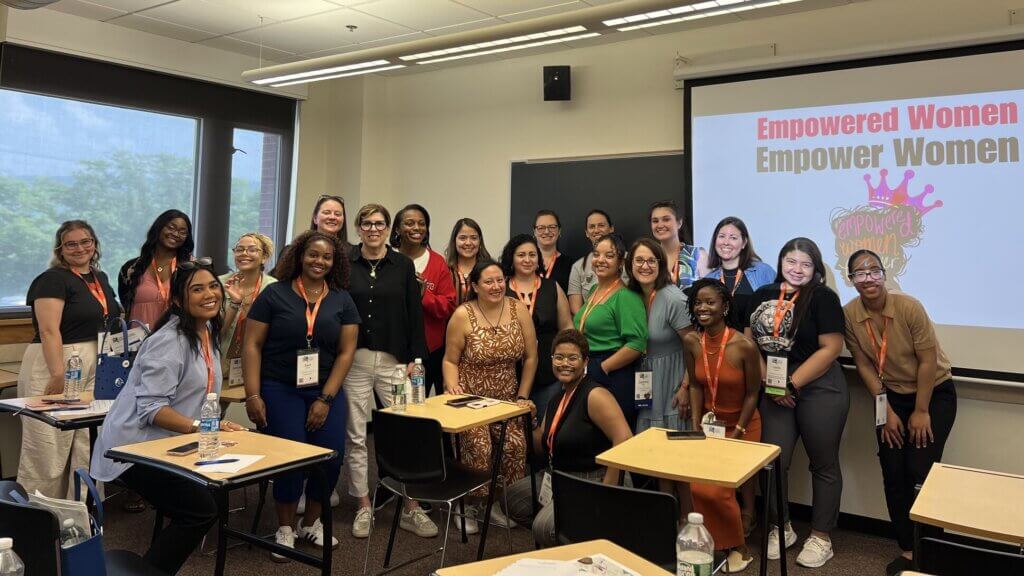 A group of women pose and smile in a classroom, some seated and some standing, with a screen behind them displaying Empowered Women Empower Women.