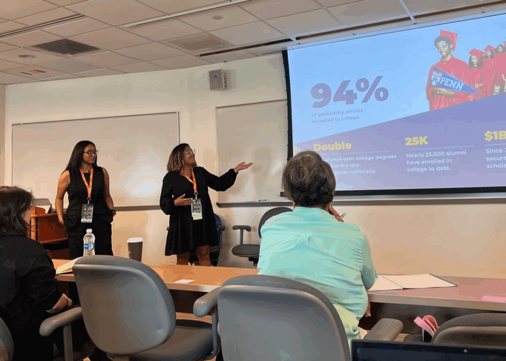 Two presenters stand at the front of a classroom, giving a presentation with a slide showing statistics about college access to an audience seated at desks.