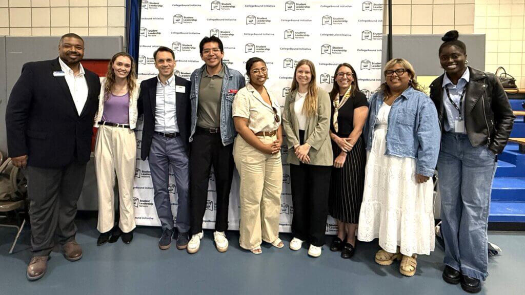 A group of nine people stands in a row, posing and smiling in front of a step-and-repeat banner inside a gymnasium