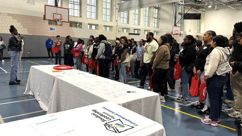 A group of people stand and listen to a speaker in a gymnasium; informational tables with materials are set up in the foreground