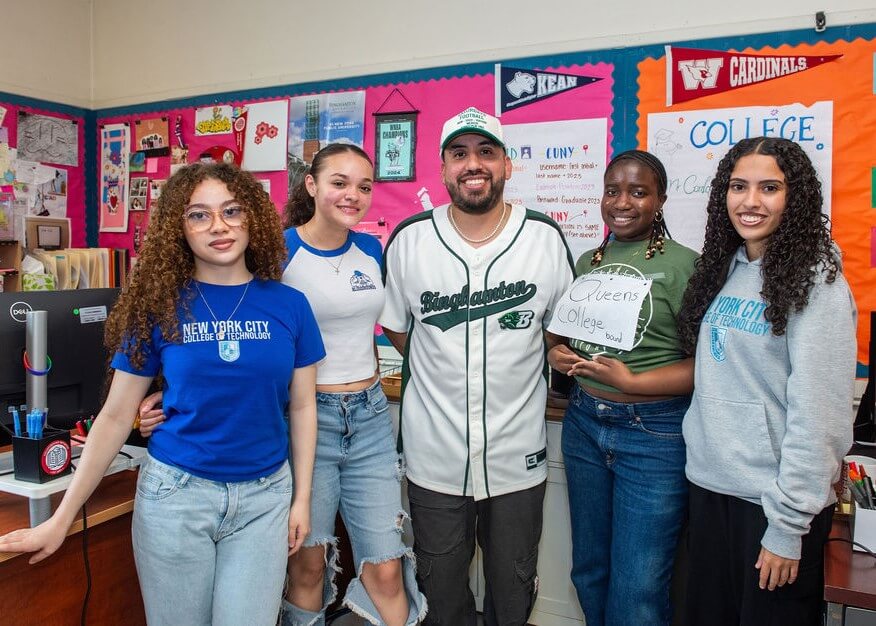 Five students stand together in a classroom decorated with college banners and posters, one holding a sign that says “Queens College.”.
