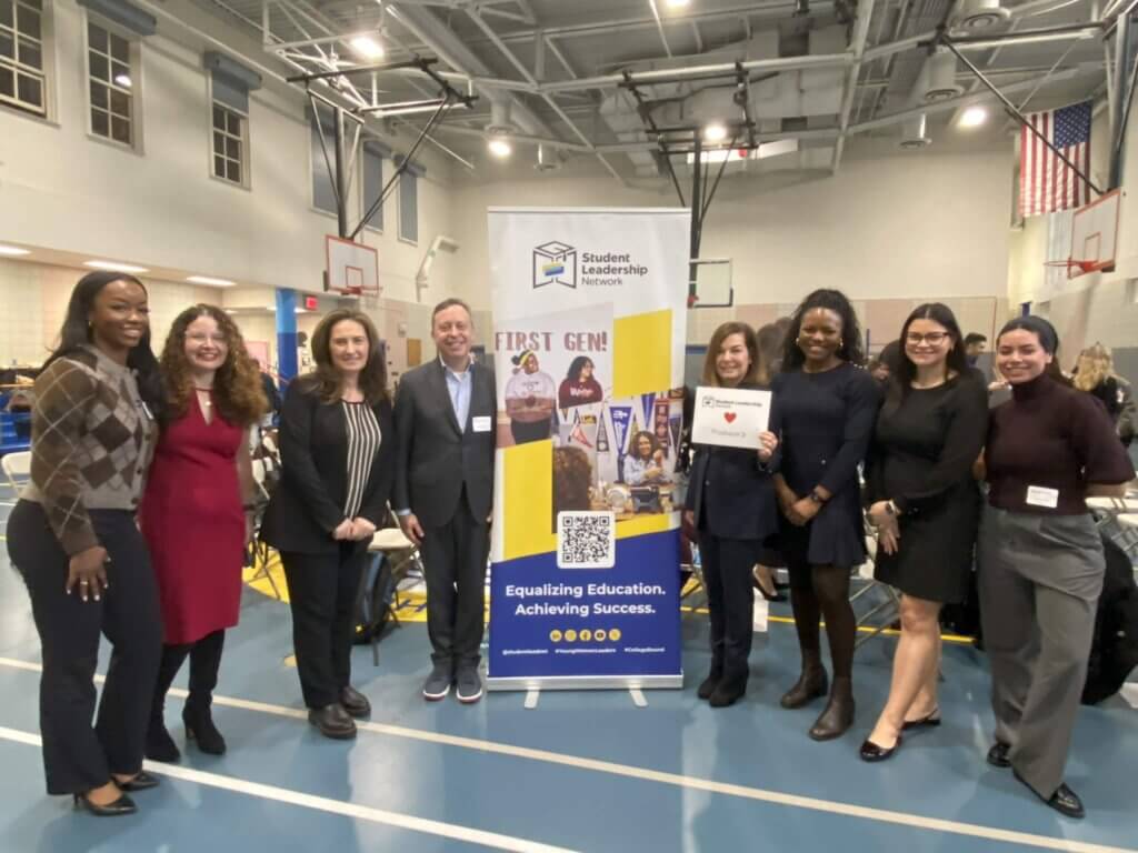 A group of eight people stands indoors next to a Student Leadership Network banner in a gymnasium, posing for a group photo.