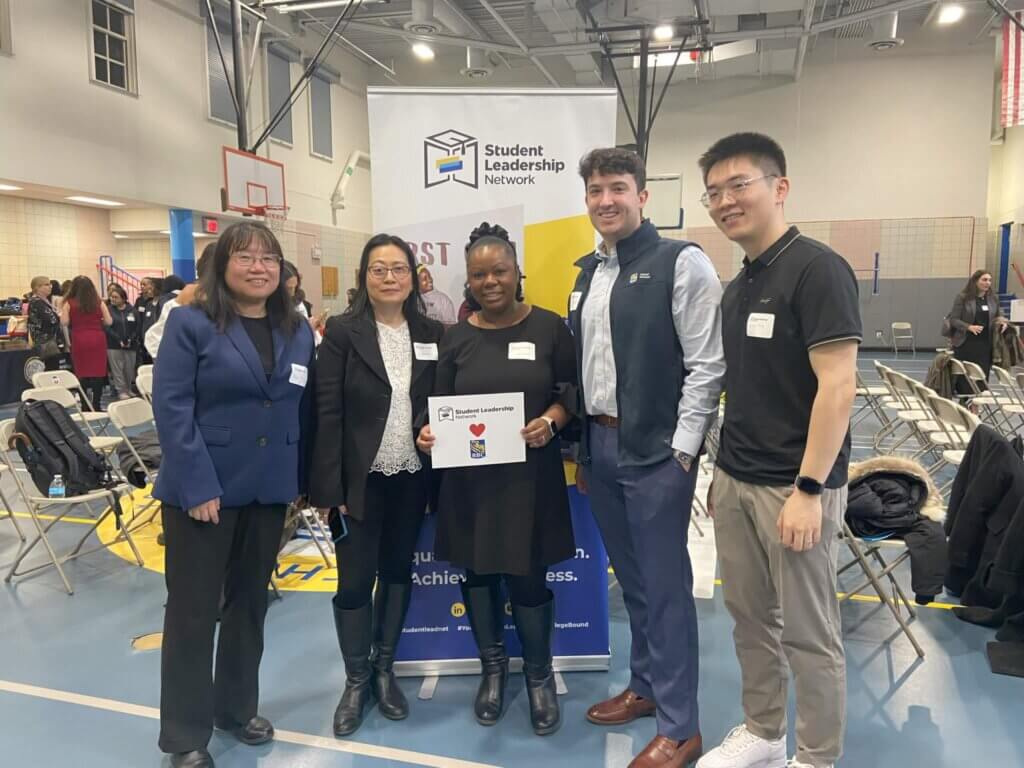 Five people stand together indoors in front of a Student Leadership Network banner, posing for a group photo at an event